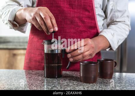 Gros plan des mains d'un homme avec un tablier rouge appuyant le café dans une presse française sur un comptoir. Mise au point sélective Banque D'Images