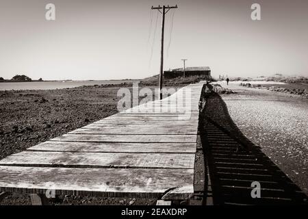 Passerelle en bois rustique menant à l'ancien hangar de côté d'eau et à la colonne de puissance avec des lignes coupées danseuses d'Ocean Beach Road à Bluff Invercargill, Nouvelle-Zélande. Banque D'Images