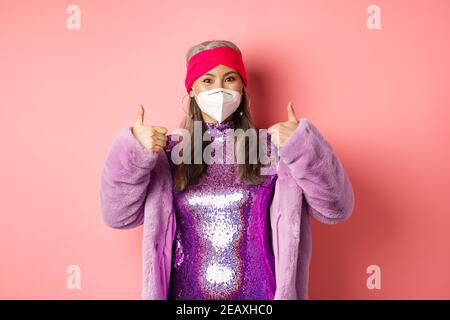 Covid-19, pandémie et mode concept. Une femme asiatique âgée gaie faisant la fête pendant le coronavirus avec un respirateur, montrant le pouce vers le haut, recommande l'usure Banque D'Images