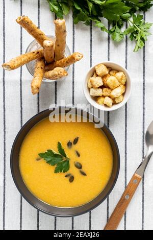 Un bol de soupe de potiron biologique crémeuse épicée maison sur fond de tissu blanc rayé avec des bâtonnets de pain, de la crème sure, du persil et des épices. Wi Banque D'Images