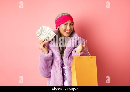 Femme asiatique riche et à la mode gaspillant de l'argent dans les magasins, tenant sac de shopping et dollars, en se faisant un plaisir à l'appareil photo, fond rose Banque D'Images