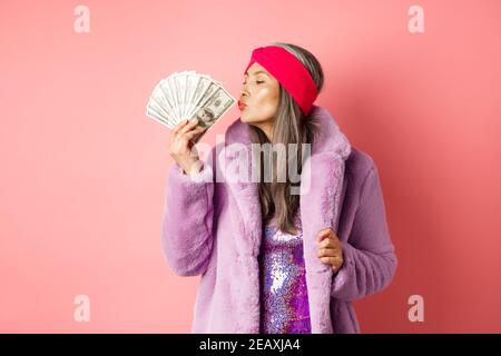 Shopping et mode. Tendance et riche femme senior baiser dollars d'argent, regardant heureux, portant manteau violet en fausse fourrure avec robe de fête Banque D'Images