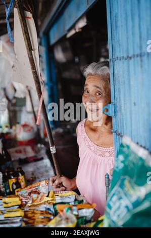 Une femme asiatique âgée vend des bonbons et des boissons gazeuses dans sa petite fenêtre, souriant à la caméra. Banque D'Images