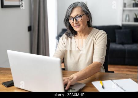 Femme sénior et confiante dans des lunettes utilisant un ordinateur portable pour travailler à distance, assise au bureau à la maison. Une femme âgée concentrée tapant des réponses à des courriels Banque D'Images