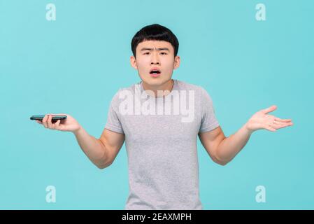 Malheureux jeune asiatique homme haussant l'épaule ayant des problèmes avec son smartphone isolé sur fond de studio bleu clair Banque D'Images