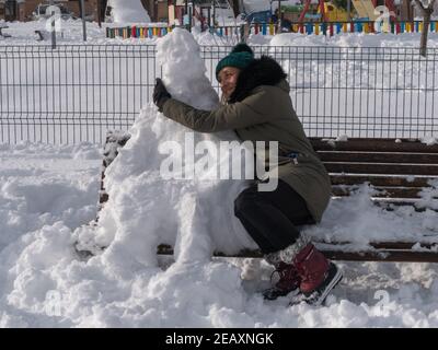 Blonde gaie d'âge moyen dans un manteau assis sur un banc embrassant un bonhomme de neige dans un parc Banque D'Images