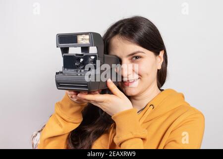 Belle dame brunette tient un appareil photo et prend des photos, souriant sur un fond blanc. Photographe féminin, cours et formation en photographie. Banque D'Images