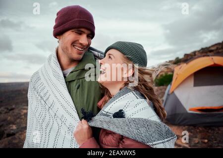 Rire jeune couple romantique dans une couverture debout ensemble à l'extérieur tente sur la montagne regardant l'un l'autre Banque D'Images