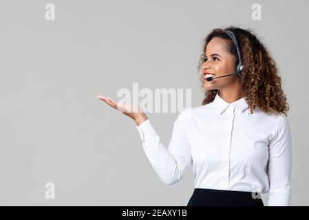 Service souriant femme afro-américaine sourit de porter des écouteurs personnel du centre d'appels avec main ouverte sur fond gris clair Banque D'Images