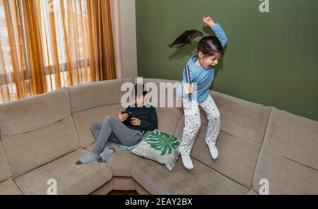 Fille sautant et jouant sur le canapé pendant que son frère regarde la tablette en position assise Banque D'Images