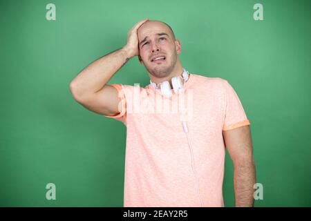 Jeune homme chauve utilisant des écouteurs sur fond vert en mettant un main sur sa tête en souriant comme il avait oublié quelque chose Banque D'Images