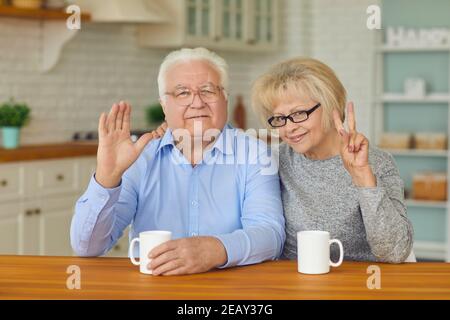 Heureux couple de personnes âgées grands-parents assis ensemble dans la cuisine, agitant et montrant le signe de paix Banque D'Images