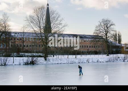 ZUTPHEN, PAYS-BAS - 10 févr. 2021: Patin à glace seul sur un lac gelé naturel l'après-midi ensoleillé avec bâtiment et tour d'église à l'arrière Banque D'Images