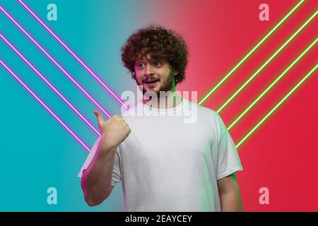 Portrait du jeune homme caucasien isolé sur fond de studio bleu-rouge dégradé dans une lumière néon avec lignes violettes-vertes. Look futuriste. Concept des émotions humaines, expression faciale, ventes, publicité. Banque D'Images