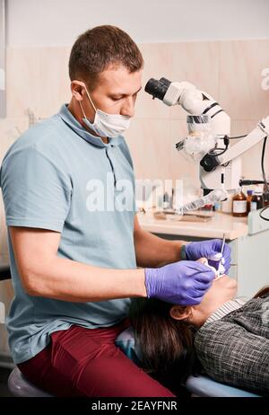 Jeune homme dentiste en masque médical utilisant un microscope de diagnostic dentaire tout en traitant les dents du patient. Femme couchée sur une chaise pendant un examen dentaire dans un centre médical moderne. Concept de la dentisterie. Banque D'Images