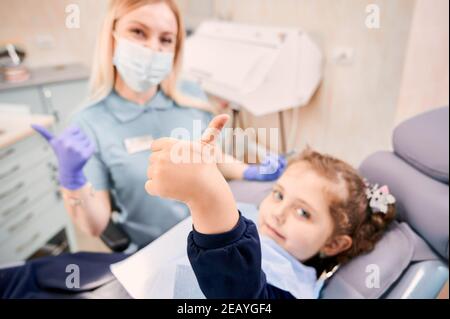 Concentrez-vous sur la main de la fille donnant les pouces vers le haut tandis que la femme dentiste dans le masque médical assis à côté de l'enfant, montrant le geste d'approbation après la procédure dentaire. Concept de la dentisterie pédiatrique et approbation des soins dentaires. Banque D'Images