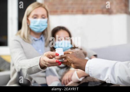 Gros plan de la photo de mère et de fille de l'adolescence brouillées dans des masques médicaux, malades à la maison. Mère d'âge moyen prenant une bouteille avec des comprimés d'elle Banque D'Images