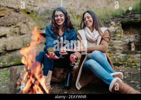 jeunes femmes heureux de boire un verre de vin rouge. Les femelles se réchauffent à côté du feu. Feu de camp, activités de plein air concept. Banque D'Images