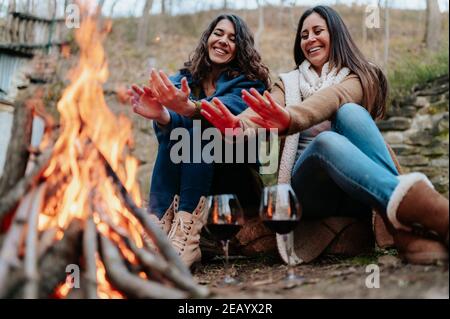 les jeunes femmes souriantes se réchauffent près du feu. Verres de vin rouge. Feu de camp, activités de plein air, détente, concept de togetherness. Banque D'Images