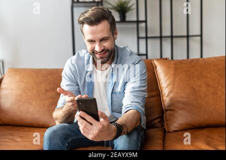 Un homme souriant portant une tenue décontractée parle par connexion vidéo sur le smartphone. Vue de face un jeune homme s'assoit sur le canapé moderne et utilise l'application mobile pour Banque D'Images