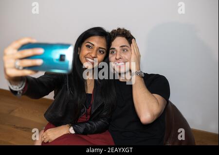 Jeune couple multiethnique prenant un autoportrait avec un smartphone à la maison assis sur un fauteuil. Banque D'Images