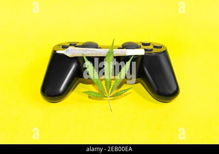 Jeux vidéo et cannabis: Contrôleur de jeu avec feuille de marijuana et grand isolat conjoint de cannabis sur fond jaune. Banque D'Images