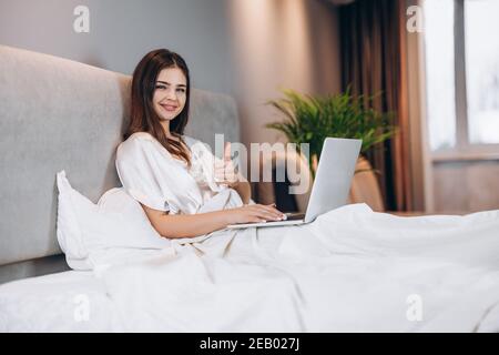 Jeune belle femme avec un ordinateur portable dans le lit. Un brunette avec un ordinateur portable regarde la caméra et montre cool. Une femme travaille à un ordinateur dans sa chambre à Banque D'Images