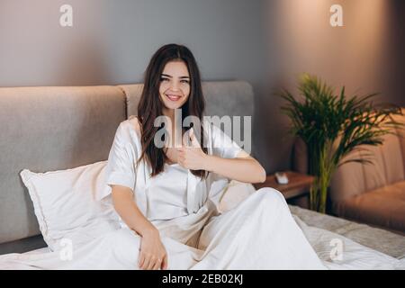 le modèle féminin en pyjama de soie blanche est très tendance. Jeune belle femme avec un sourire sur son visage assis sur le lit montre bien Banque D'Images