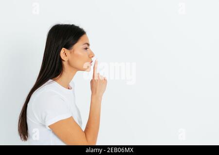 Vue latérale jeune femme montrant le geste de la ruche tenant l'index près des lèvres tout en se tenant sur fond blanc studio avec l'espace de copie. Banque D'Images