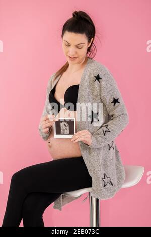 Une échographie obstétricienne imprimée tenue devant le ventre d'une femme enceinte sur fond rose. Mise au point sélective. Concept de grossesse. Banque D'Images