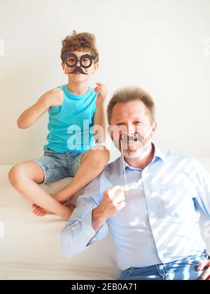 Le père et son petit fils mignon ont mis sur la barbe en papier et ont le sourire. La famille se prépare à célébrer le jour des Fools d'avril. Banque D'Images