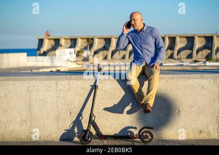 un homme mûr parle au téléphone. Il est assis et avec un scooter électrique. Photo horizontale prise à l'extérieur avec la mer en arrière-plan. Banque D'Images