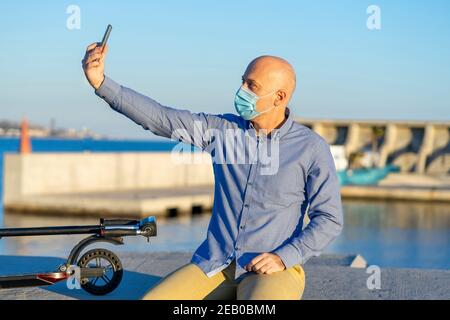 Un homme mûr prend un selfie avec son téléphone portable, portant un masque. Il est assis et a un scooter électrique. En arrière-plan, vous pouvez voir la mer. Banque D'Images