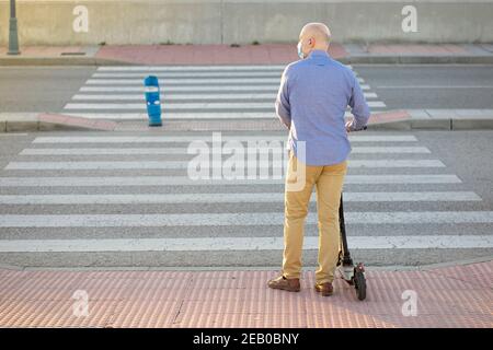 un homme avec un scooter électrique attend de traverser le passage piéton. Il porte une chemise bleue et un pantalon jaune. Son dos à la caméra. Phot horizontal Banque D'Images
