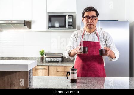 Homme en tablier rouge et verres donnant deux tasses de café filtre à la presse française à un comptoir de cuisine. Banque D'Images