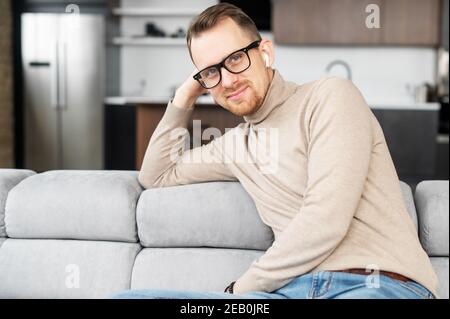 Un portrait de jeune homme beau portant des lunettes élégantes et des vêtements décontractés élégants, un homme attrayant assis sur le canapé confortable à la maison, regarde Banque D'Images