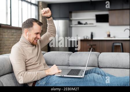 Un jeune homme regarde un match sportif sur un ordinateur portable, hurle avec joie parce que son équipe est gagnante. Concept de victoire. Un jeune homme heureux a reçu de bonnes nouvelles sur Banque D'Images