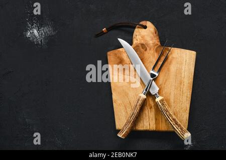 Vue en hauteur de la planche à découper en bois avec couteau à steak et fourche sur fond noir de merde Banque D'Images