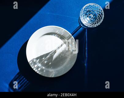 Table avec plaque blanche vide et verre cristal isolé sur fond bleu foncé, ombres sombres et lumière du soleil se reflétant à travers le verre à vin Banque D'Images