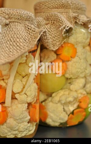 Concept de deux pots de cornichons traditionnels de saison recouverts de tissu de sac, rempli de tomates vertes crues de couleur, de carottes d'orange et de chou-fleur blanc Banque D'Images