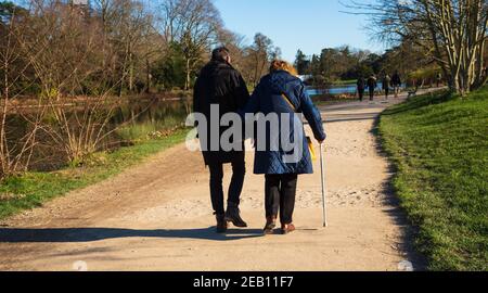 Femme senior avec bâton de marche et son fils mature faisant promenade dans le parc près du lac Daumesnil (Paris, France) en rare journée ensoleillée d'hiver. Vue arrière. El Banque D'Images