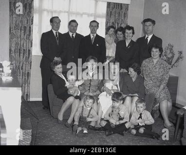 dans les années 1950, historique, une grande famille se réunit dans la salle d'entrée d'une maison pour une photo de groupe, avec quatre générations toutes ensemble, peut-être après avoir baptisé le nouveau bébé tenu par sa grand-mère sur le canapé. Un petit garçon est assis tenant son ours en peluche. Banque D'Images