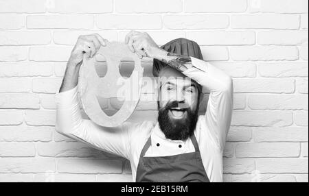 Homme avec barbe tient la pâte roulée avec smiley coupé sur fond de mur de brique blanche. Faites cuire avec un visage excités dans des pièces d'uniforme bordeaux avec de la pâte à pizza. Concept du processus de cuisson. Le chef prépare de la pâte. Banque D'Images