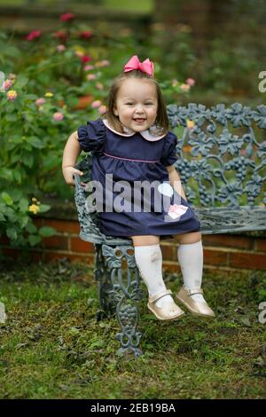 Une petite fille avec une robe marine et rose bow est assis sur un banc en fer d'époque Banque D'Images