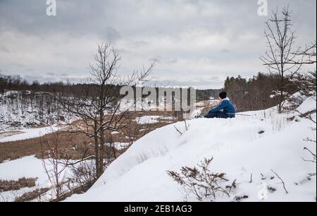 Adolescent assis au sommet d'une colline enneigée surplombant la vallée le jour d'hiver. Banque D'Images