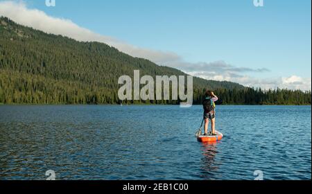 Vue arrière d'une femme méconnaissable avec pédalo à dos sur l'eau du lac ondulée près de la pente de montagne avec forêt verte en été à Whistler, CAN Banque D'Images