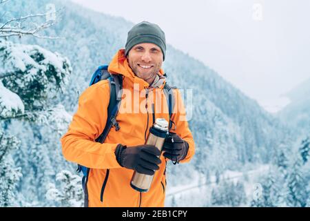 Laughing Man vêtu d'une veste Softshell orange vif avec une thermos pour boissons chaudes regardant l'appareil photo pendant qu'il trekking route des montagnes d'hiver. Actif Banque D'Images