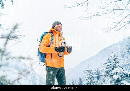 Smiling Man under Snowfall habillé blouson Softshell orange vif avec une thermos boisson chaude en trekking route des montagnes d'hiver. Peop actif Banque D'Images