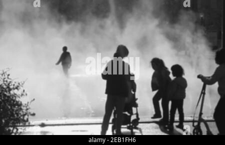 Photo floue des enfants jouant dans la brume d'eau de fontaine et de leur mère avec bébé poussette les regardant. Place des Halles, Paris, France. Noir a Banque D'Images