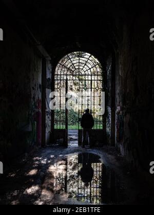 Silhouette d'un homme observant le monde extérieur regardant de la grille d'une porte et son image sont reflétées dans une flaque à l'intérieur d'un bâtiment abandonné Banque D'Images